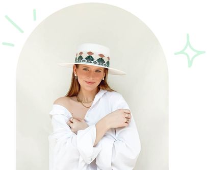 Brittany Merrill wearing white oversized shirt, white wide-brimmed hat with boho patterned trim and gold chain jewelry